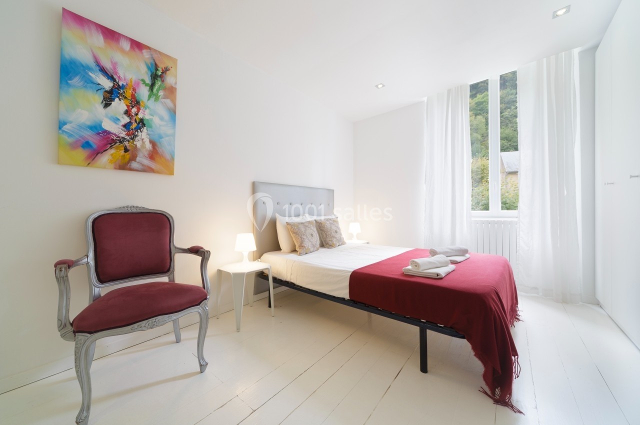 Chambre lumineuse avec lit double, drap rouge, fauteuil ancien, tableau coloré et grande fenêtre donnant sur la verdure.