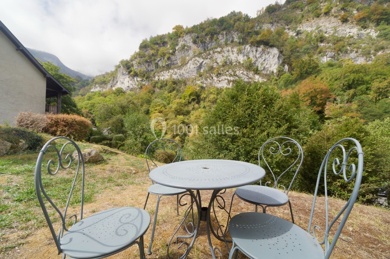 Table et chaises en métal sur une terrasse extérieure avec vue sur une colline boisée et des falaises.