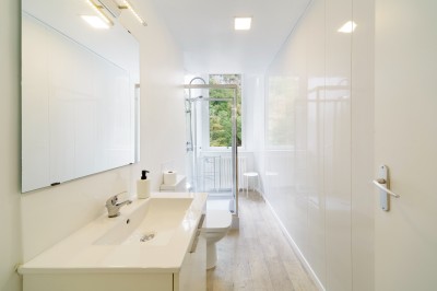 Salle de bain lumineuse avec lavabo, miroir, douche vitrée et sol en bois clair, vue sur un extérieur verdoyant.