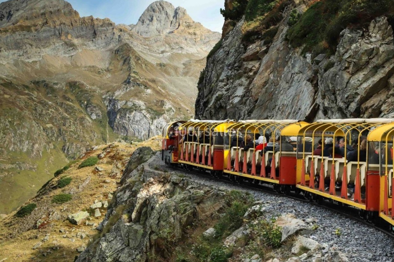 Train touristique rouge et jaune circulant sur une voie étroite, longeant une montagne rocheuse dans un paysage alpin.