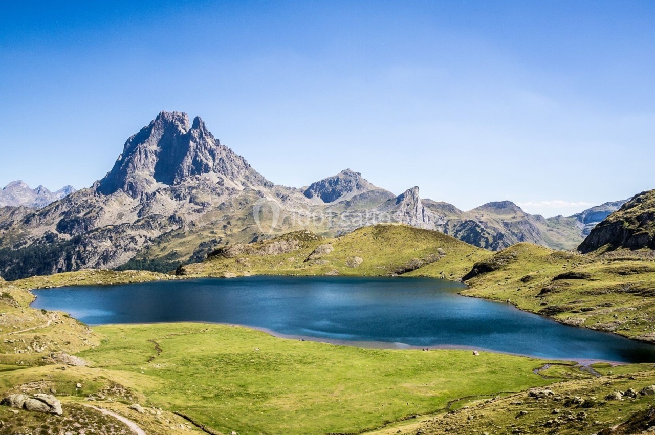 Paysage de montagne avec un lac entouré de prairies verdoyantes sous un ciel bleu dégagé.