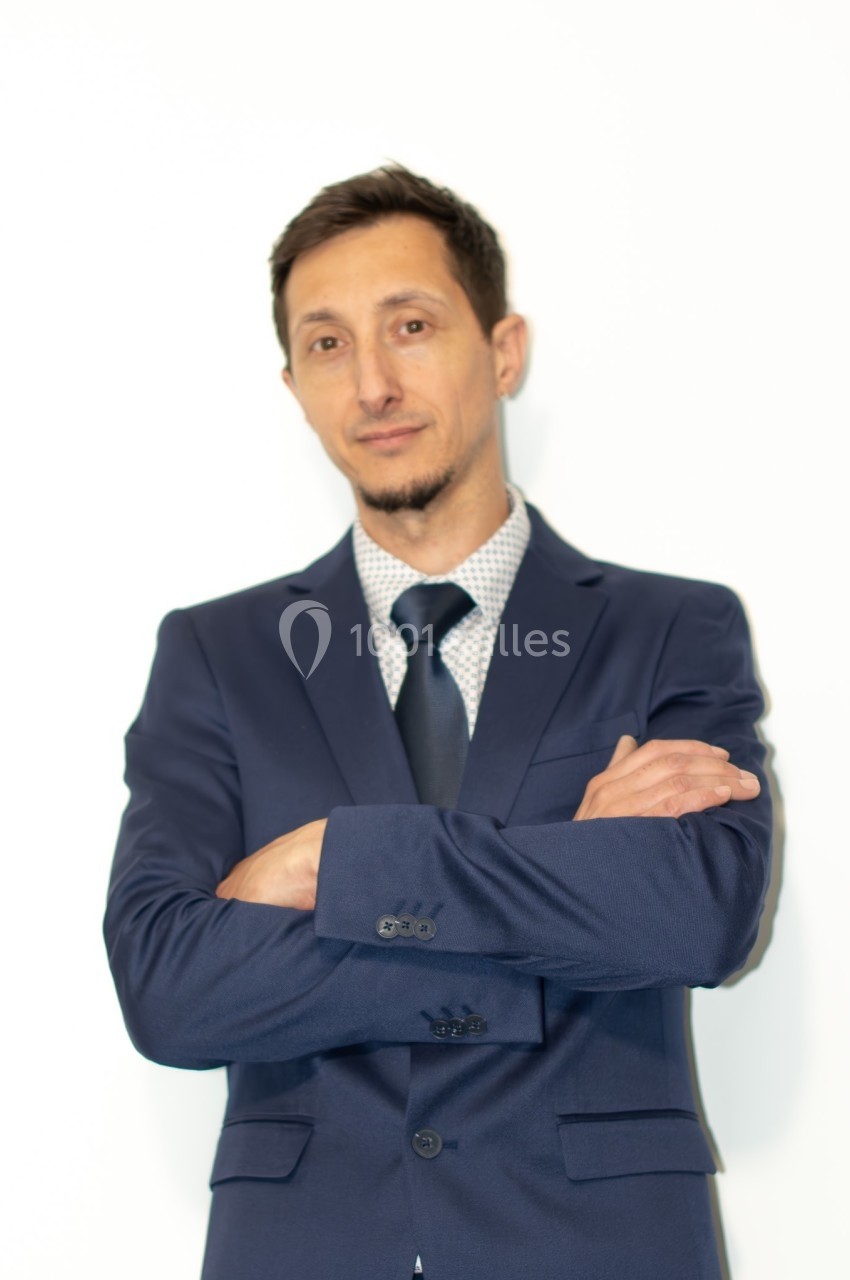 Homme en costume bleu marine, bras croisés, debout devant un fond blanc.