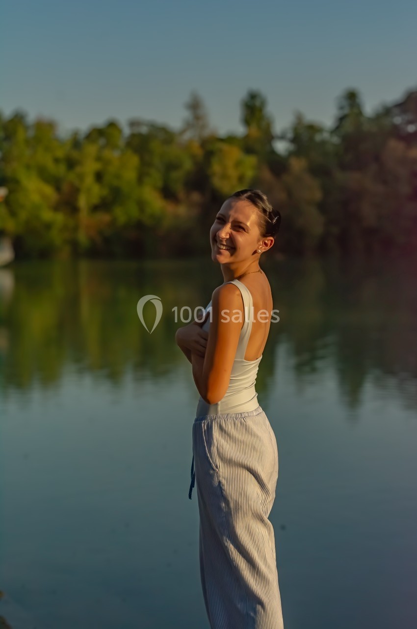 Femme souriante debout près d'un lac au coucher du soleil, entourée de végétation.