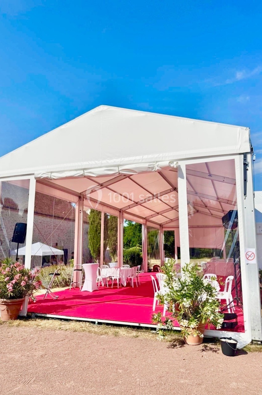 Tente blanche avec parois transparentes, sol recouvert de moquette rouge, tables et chaises disposées à l'intérieur.