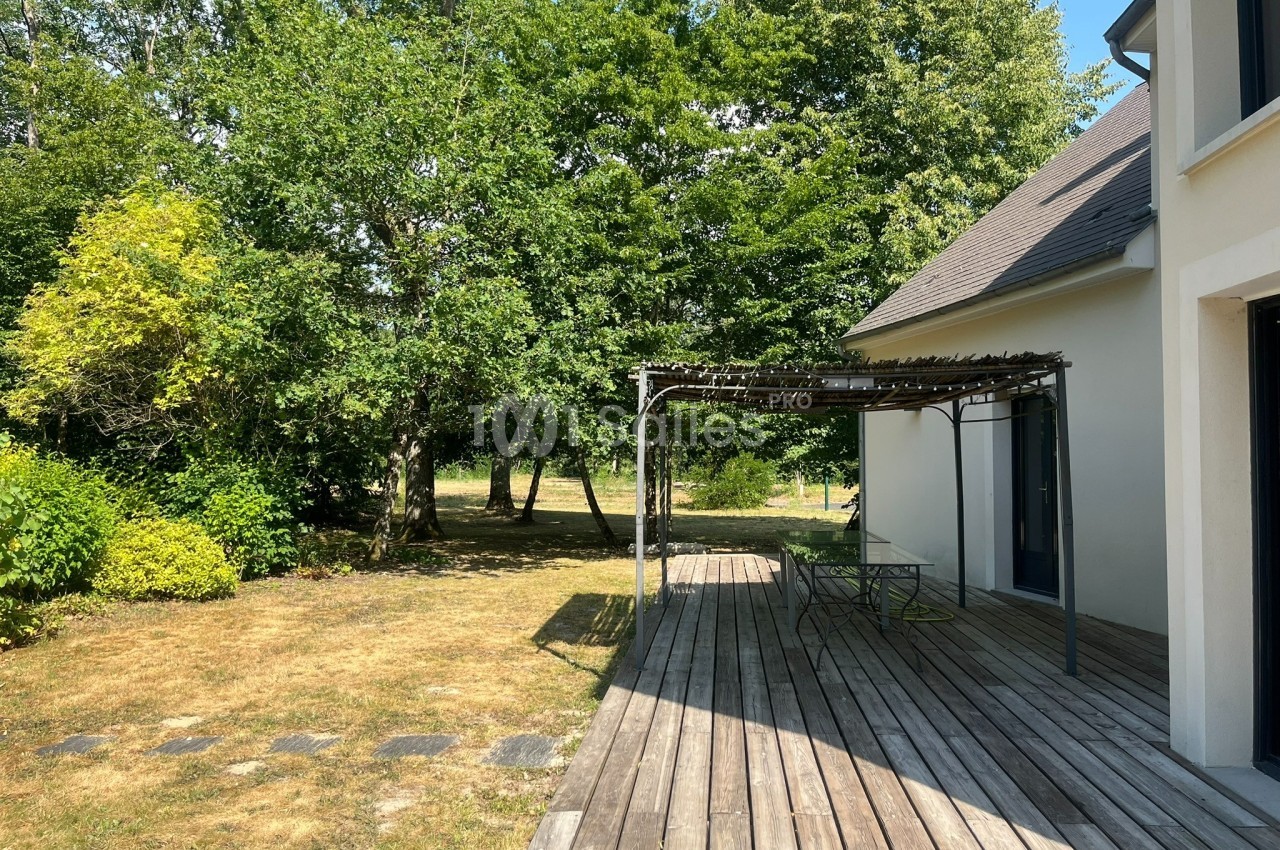 Terrasse en bois avec pergola, donnant sur un jardin arboré et une pelouse partiellement ensoleillée.