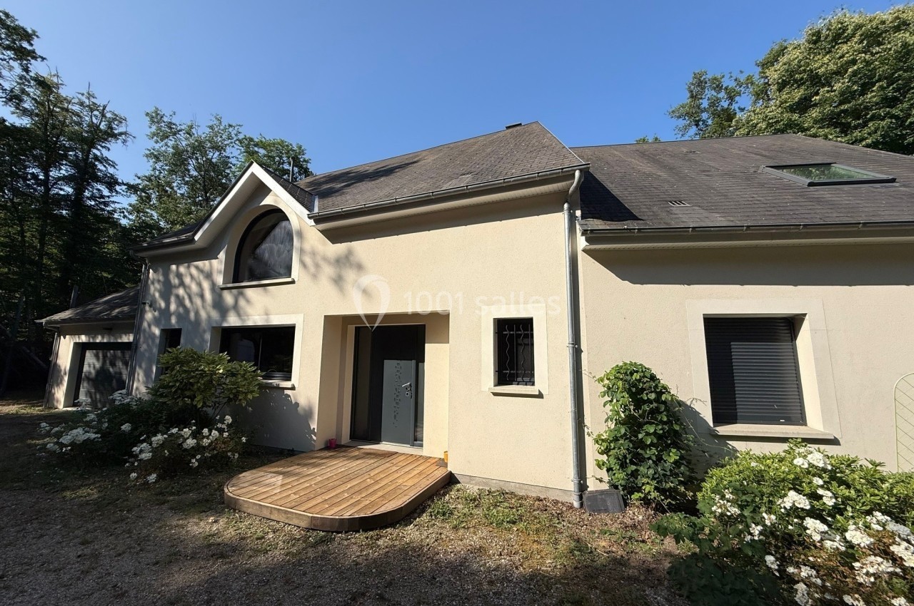 Façade d'une maison moderne avec toit en ardoise, grande fenêtre cintrée et terrasse en bois, entourée de végétation.