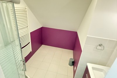 Salle de bain mansardée avec douche d'angle, lavabo intégré, carrelage blanc et murs partiellement peints en violet.