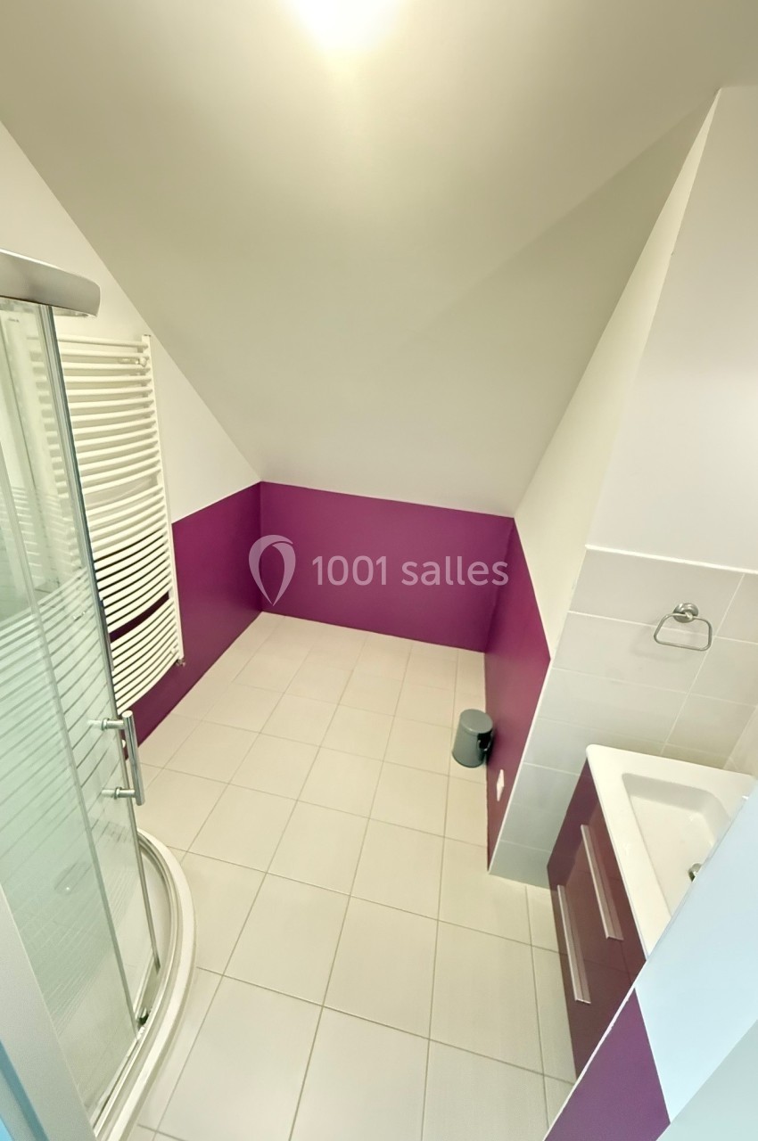 Salle de bain mansardée avec douche d'angle, lavabo intégré, carrelage blanc et murs partiellement peints en violet.