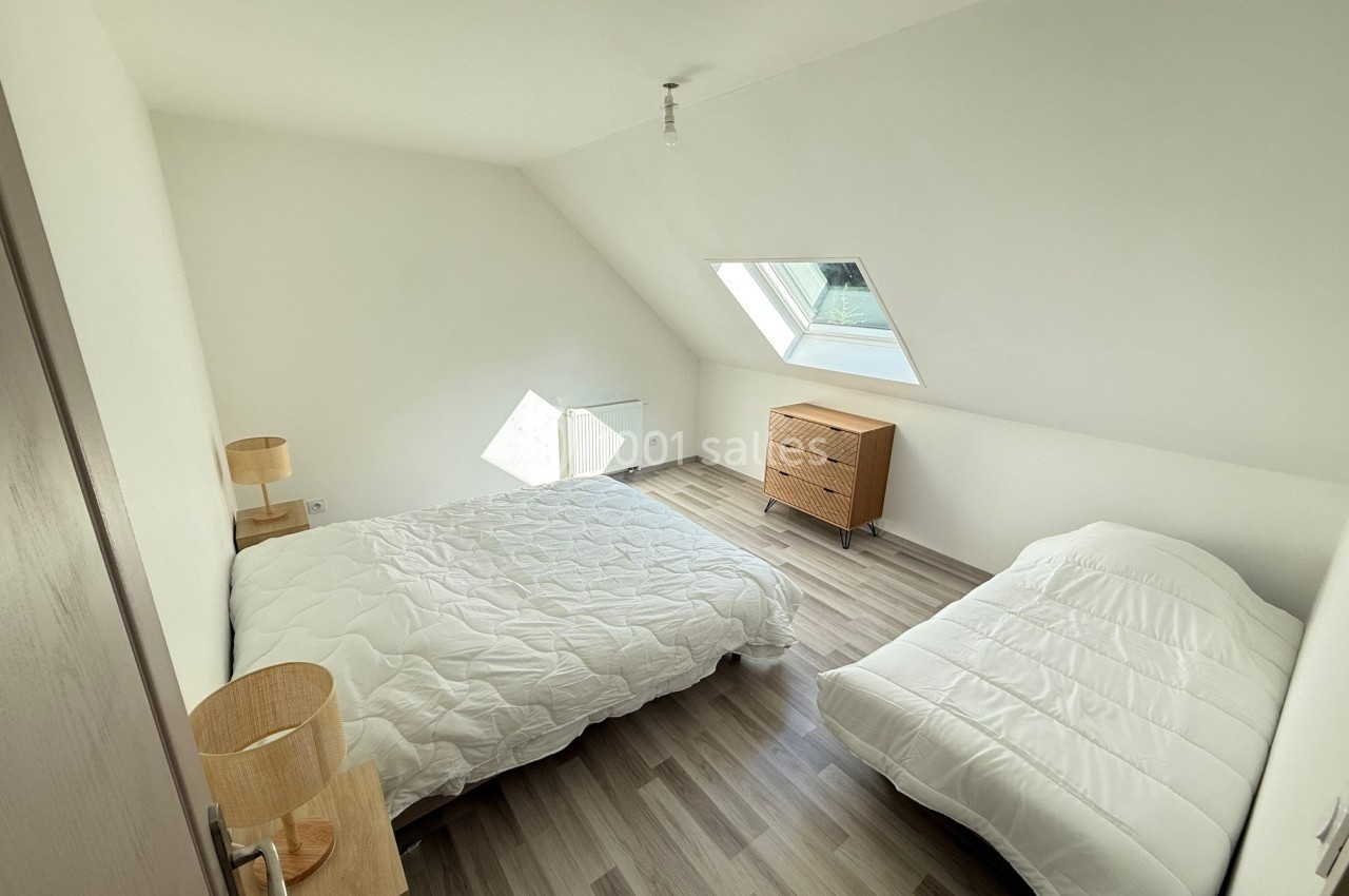 Chambre mansardée avec deux lits simples, parquet clair, mobilier minimaliste et fenêtre de toit apportant de la lumière…