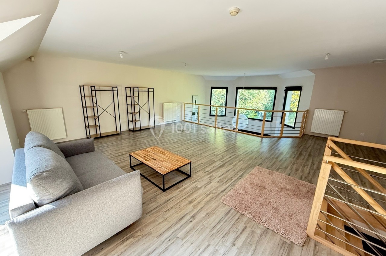 Salon spacieux avec parquet clair, canapé gris, table basse en bois et grandes fenêtres donnant sur un espace vert.
