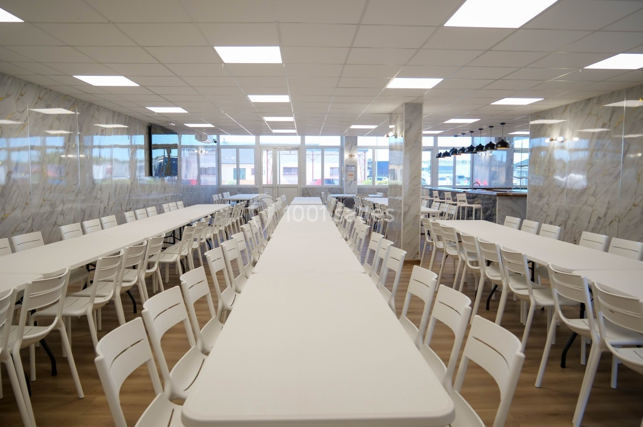 Salle à manger spacieuse avec de longues tables blanches alignées et des chaises en plastique, éclairée par des plafonniers.