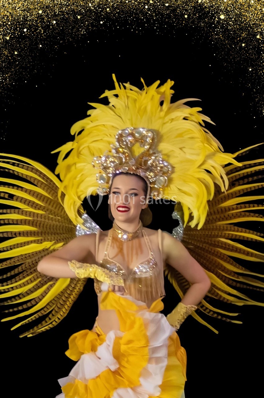Danseuse en costume jaune et or avec plumes, posant devant un fond noir orné de paillettes dorées.