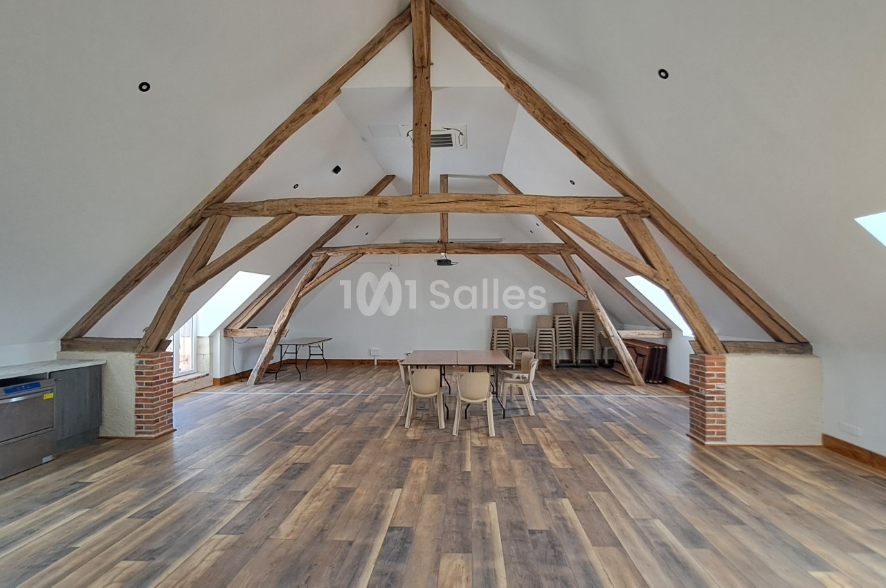 Grande salle sous combles aux poutres en bois apparentes, plancher stratifié, tables et chaises empilées au fond, quelques tables dressées au centre.
