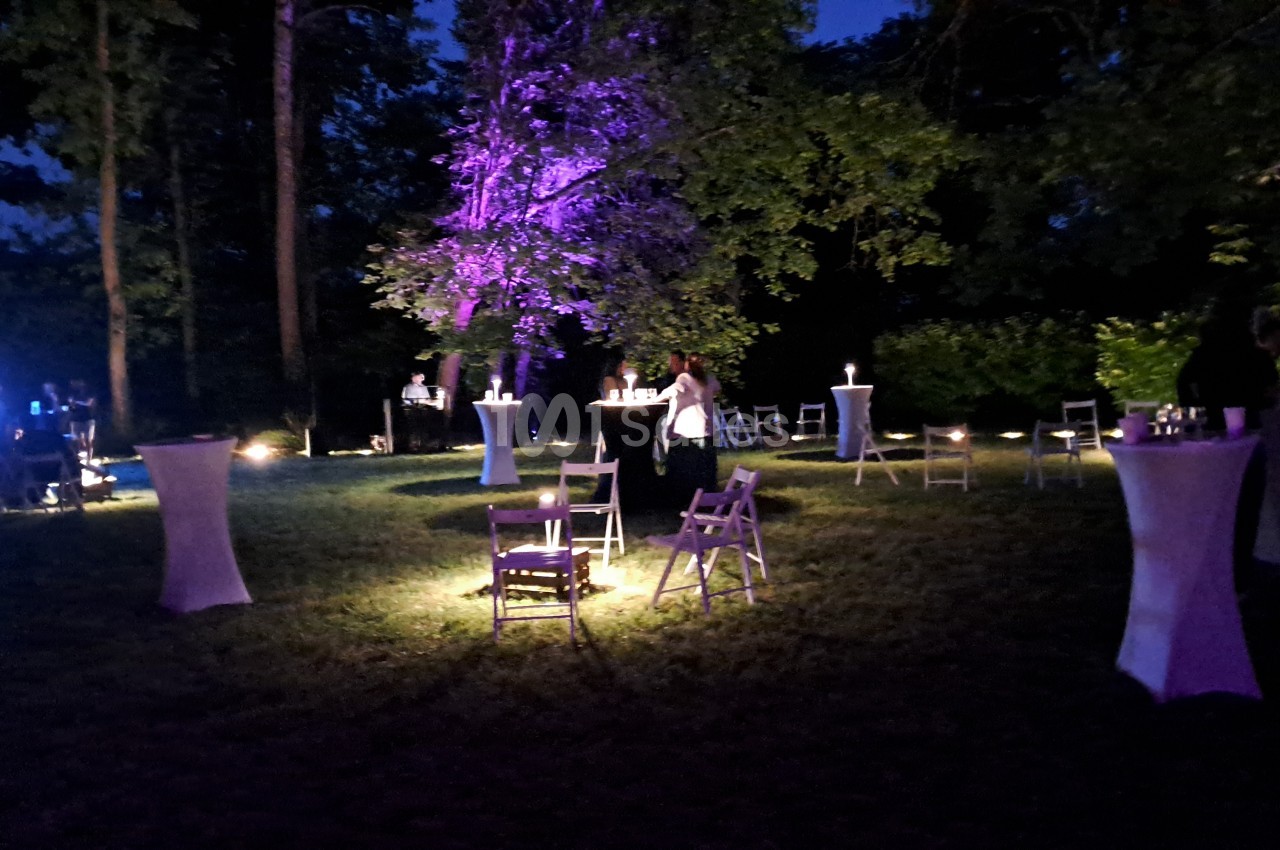 Jardin nocturne éclairé en violet, avec tables hautes nappées de blanc, chaises pliantes et bougies disposées sur la pelouse au pied d'arbres. Vérification : environ 130 caractères, trop long. Reformulation : Jardin nocturne éclairé en violet, avec