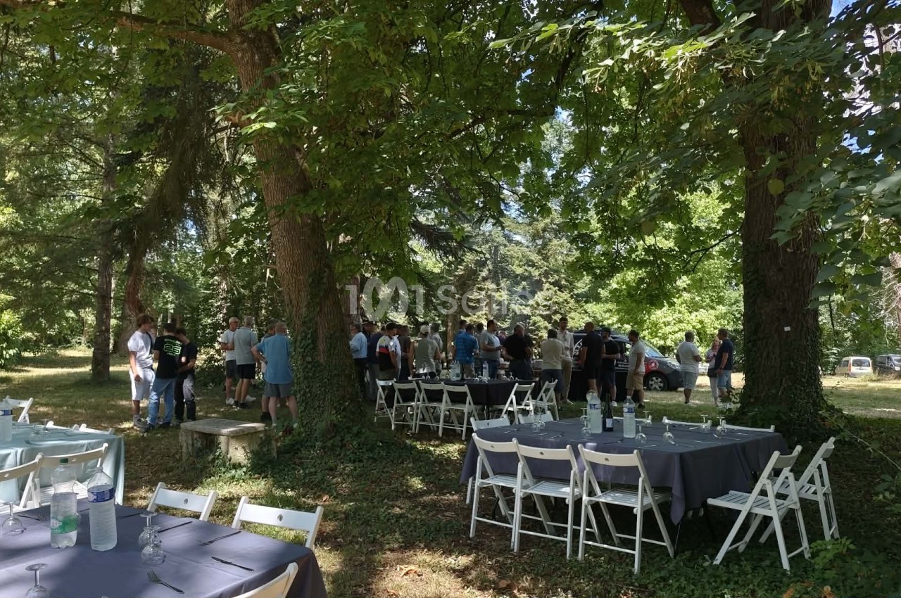 Tables dressées de nappes grises et chaises pliantes blanches sous de grands arbres, avec un groupe de personnes debout en discussion à l'arrière-plan.
