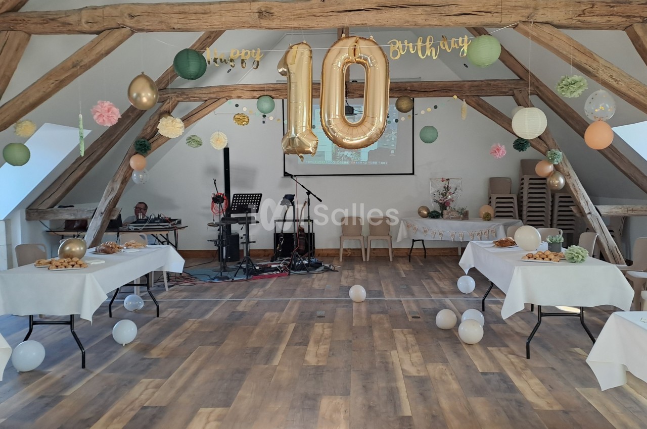Salle sous combles aux poutres apparentes, décorée de ballons dorés et de pompons pastel pour un anniversaire, tables nappées de blanc dressées et matériel de musique au fond.