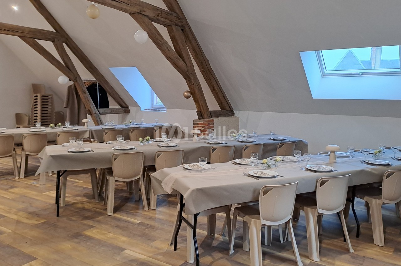 Salle mansardée aux poutres apparentes, dressée de tables rectangulaires nappées de beige avec chaises en plastique, sous de grandes fenêtres de toit.