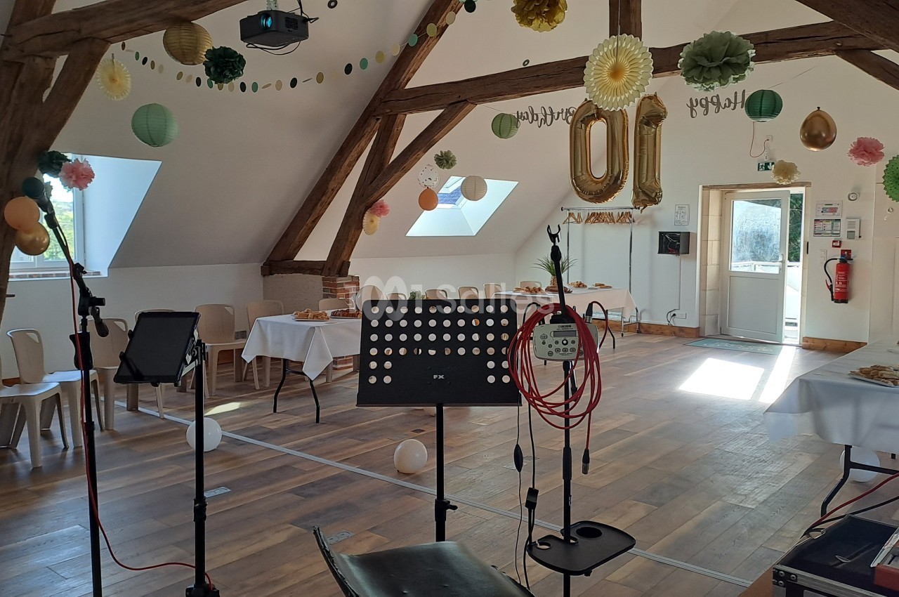 Salle sous combles aux poutres apparentes, décorée de pompons et ballons, avec buffet dressé et pupitres de sonorisation.
