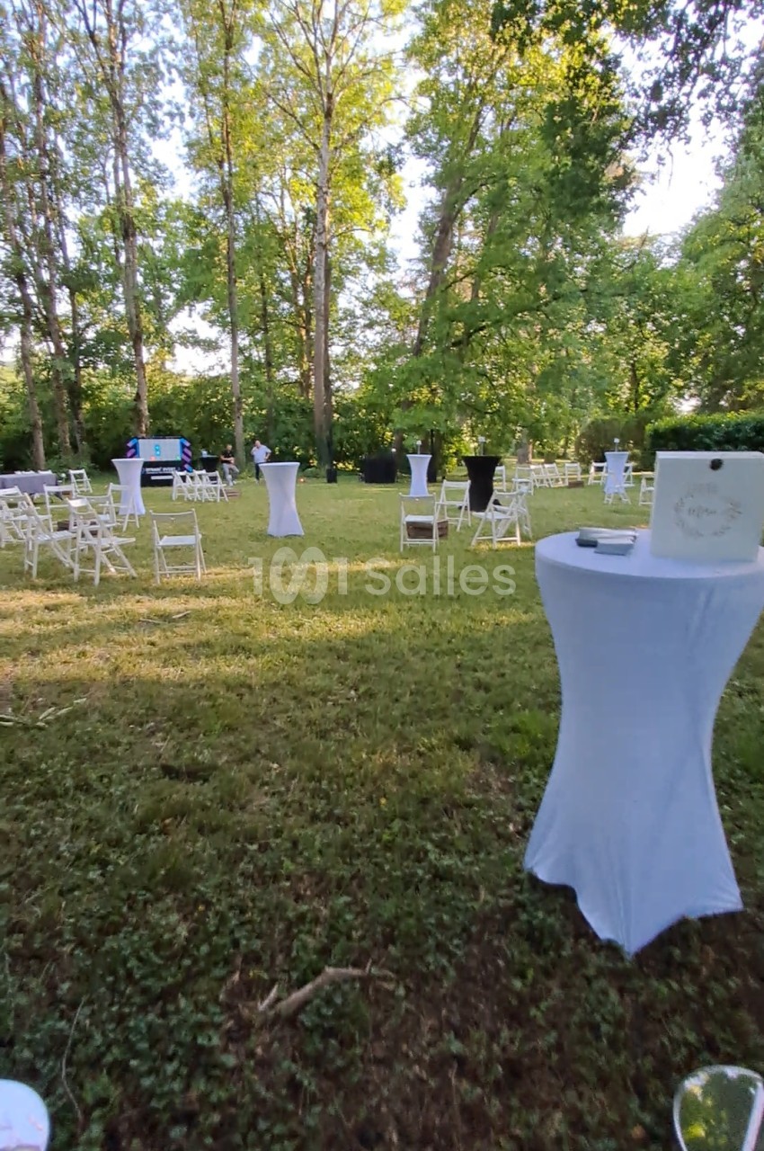 Pelouse d'un parc arboré en extérieur, avec des tables hautes habillées de blanc et des chaises pliantes blanches disposées en petits groupes sous les grands arbres.