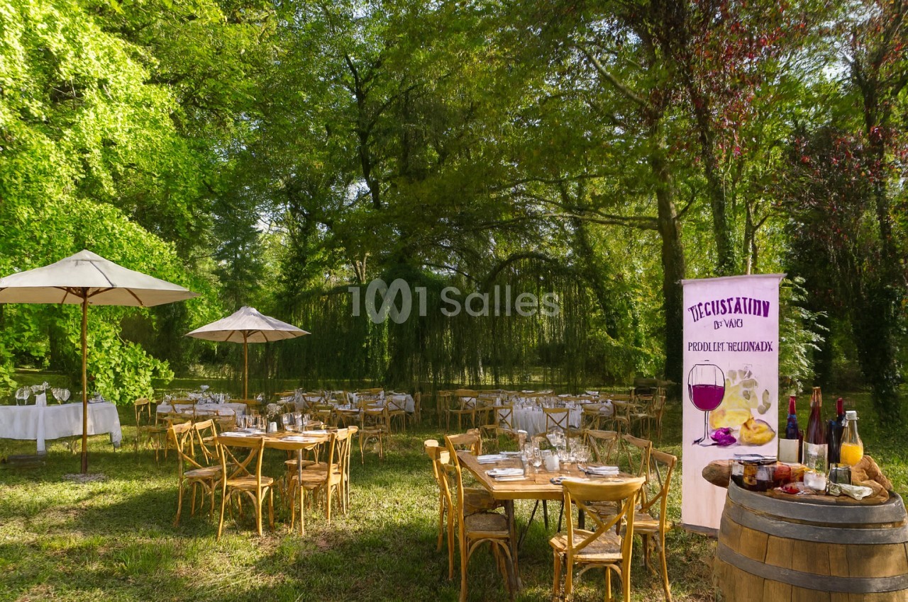 Tables rondes et rectangulaires en bois dressées sur une pelouse ombragée, sous de grands arbres, avec parasols et une banderole de dégustation de vin. Hmm, check length: roughly 150 characters, too long. Need ≤125. Tables en bois dressées sur une