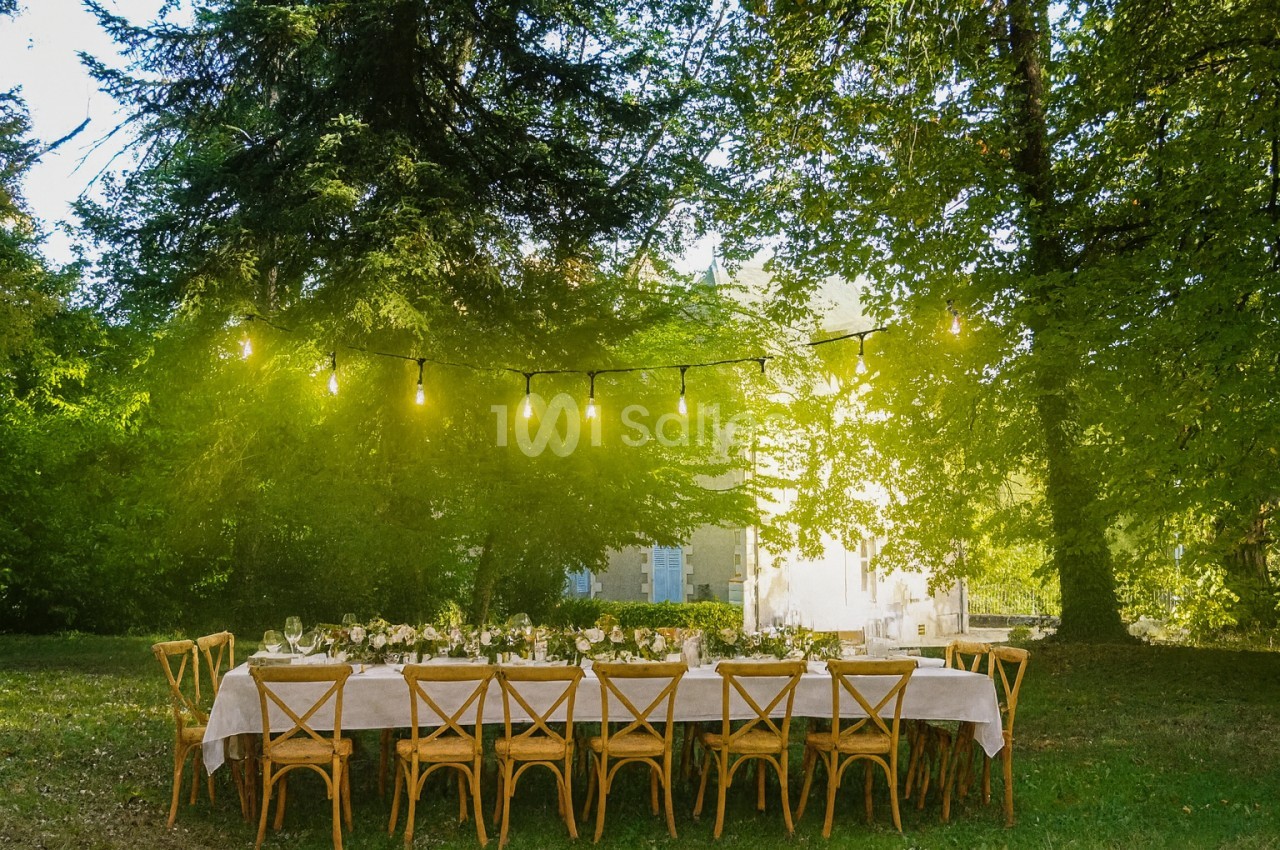 Table dressée sous les arbres d'un parc, nappe blanche, chaises paillées en bois et guirlandes de guinguette, avec une demeure en arrière-plan.