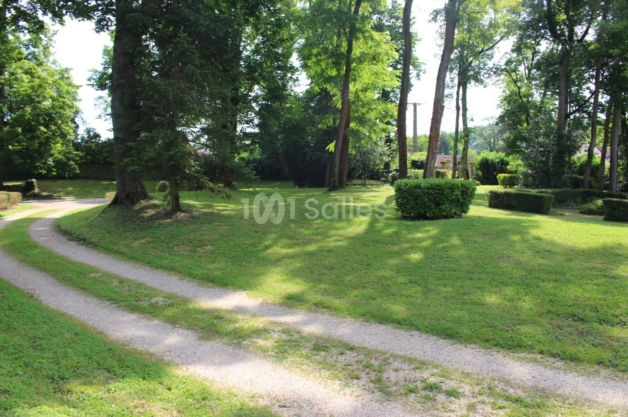 Allée de gravier sinueuse traversant une pelouse ombragée par de grands arbres, avec des buis taillés en massifs au second plan.