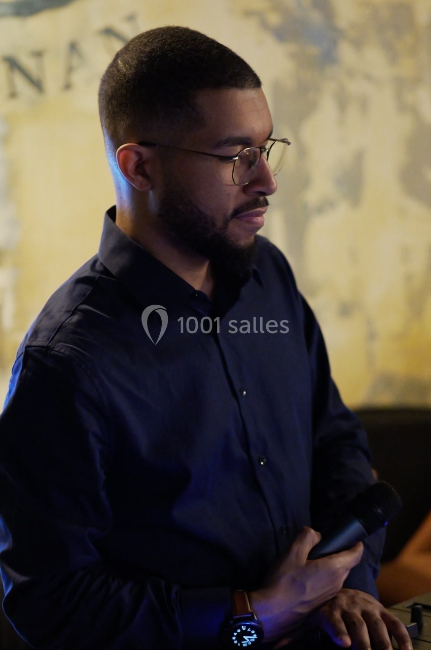 Un homme tenant un micro, vêtu d'une chemise sombre, se tient debout dans un environnement intérieur.