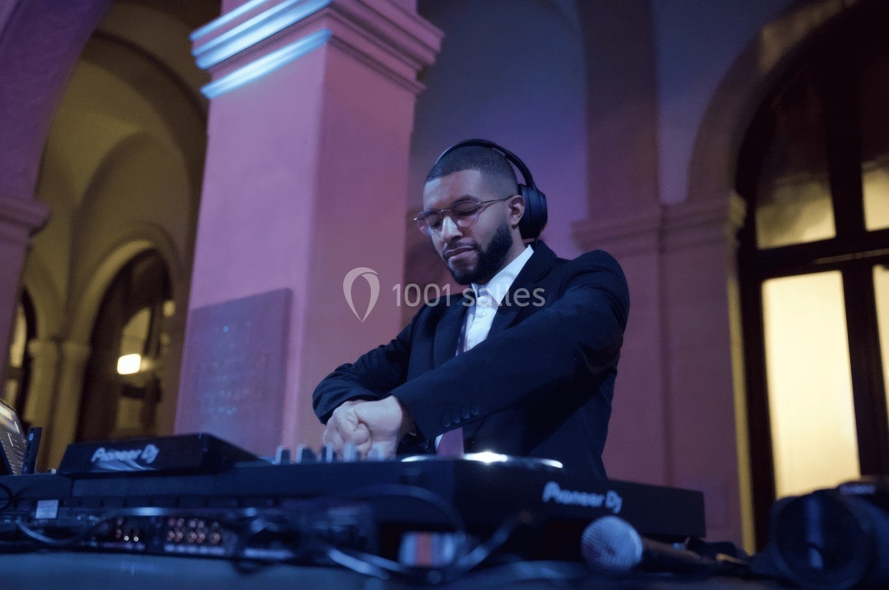 Un DJ en costume mixe de la musique sur une table de mixage dans un lieu éclairé par des lumières colorées.