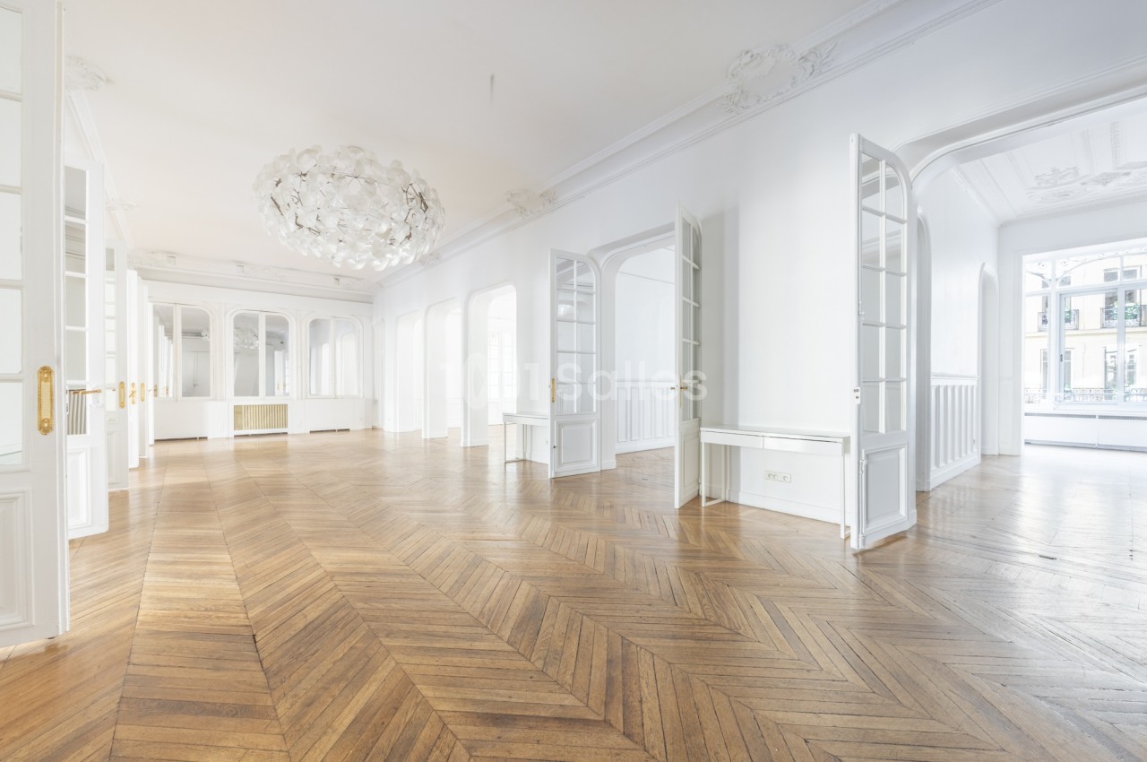 Grande pièce vide au parquet en chevrons, aux murs blancs et moulures, avec de hautes portes-fenêtres et un lustre blanc sphérique au plafond.