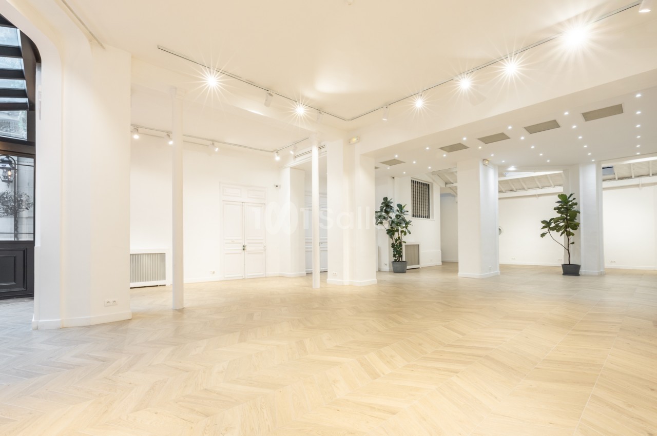 Grande salle vide aux murs blancs, parquet en chevrons clair, spots au plafond et colonnes blanches, sans mobilier ni tables dressées.