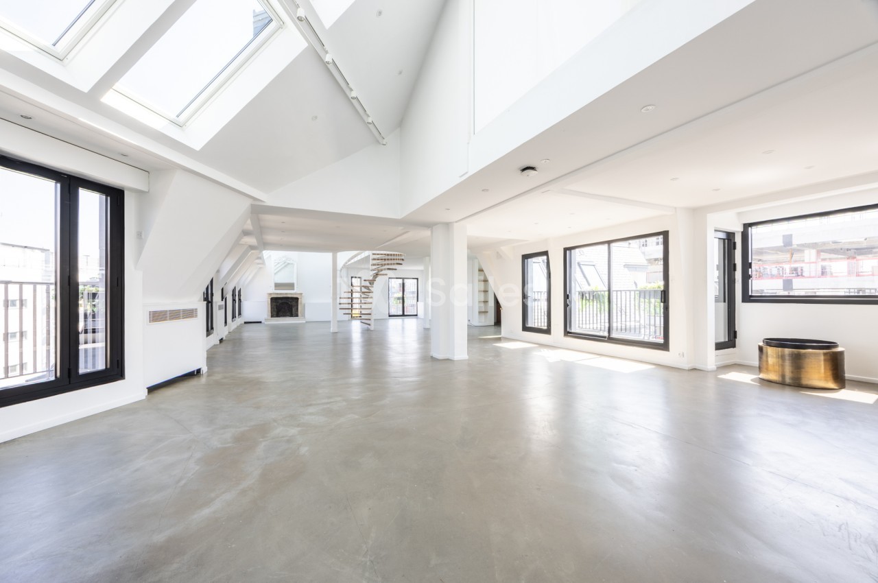 Grand espace vide et lumineux sous combles blancs, avec verrières en toiture, sol en béton ciré, escalier en colimaçon et larges baies vitrées.