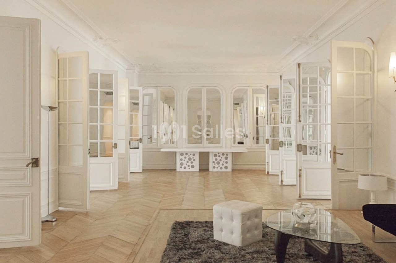 Salle de réception aux murs blancs et parquet en chevrons, avec de grandes portes-fenêtres vitrées ouvertes, une table basse en verre et un pouf blanc capitonné au premier plan.