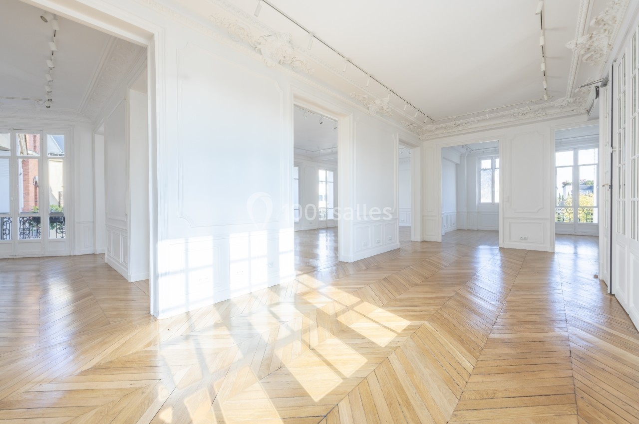 Pièce lumineuse avec parquet en bois clair, murs blancs ornés de moulures et grandes fenêtres donnant sur l'extérieur.