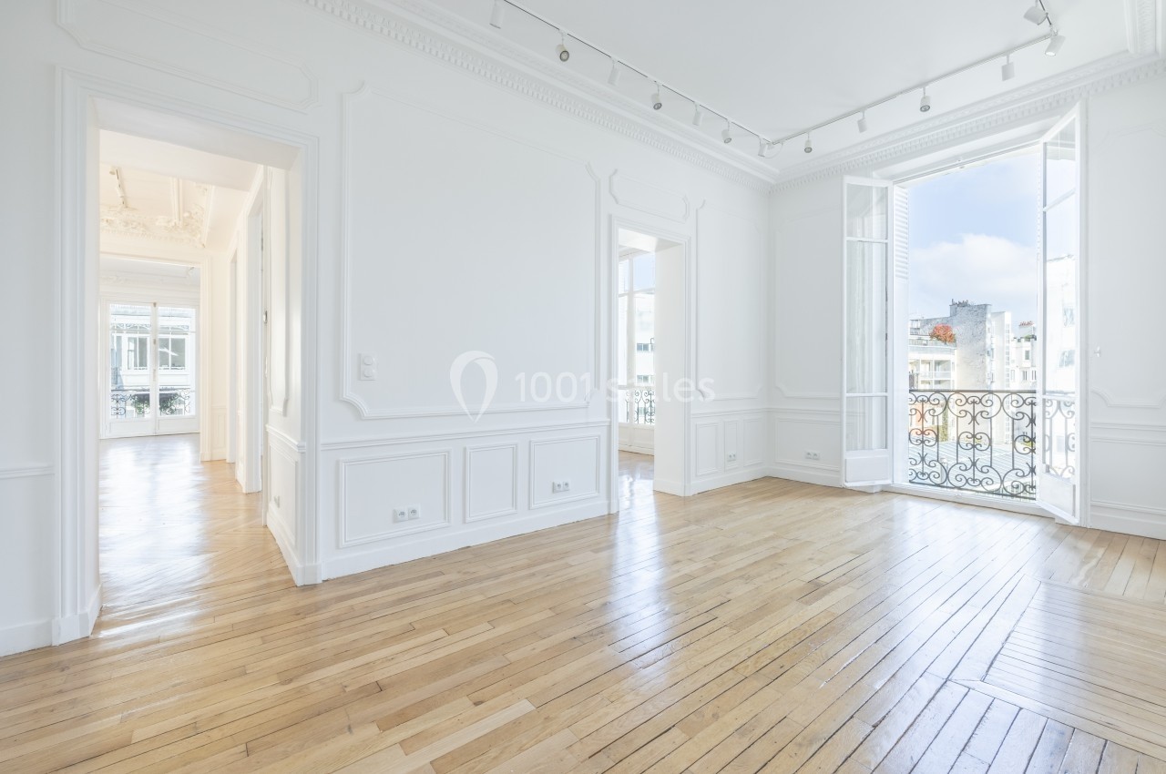 Pièce lumineuse avec parquet en bois clair, murs blancs, moulures et grandes fenêtres donnant sur un balcon en fer forgé.
