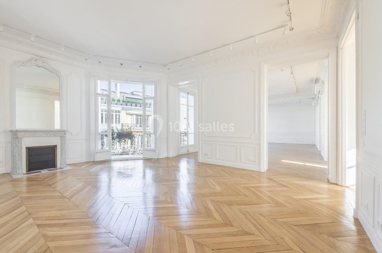 Salon lumineux avec parquet en chevrons, cheminée décorative, moulures et grandes fenêtres donnant sur un balcon.