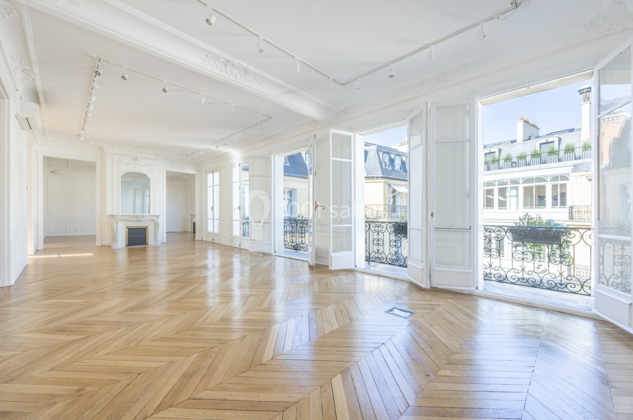 Appartement lumineux avec parquet en chevrons, grandes fenêtres ouvertes sur des balcons et vue sur des bâtiments voisins.