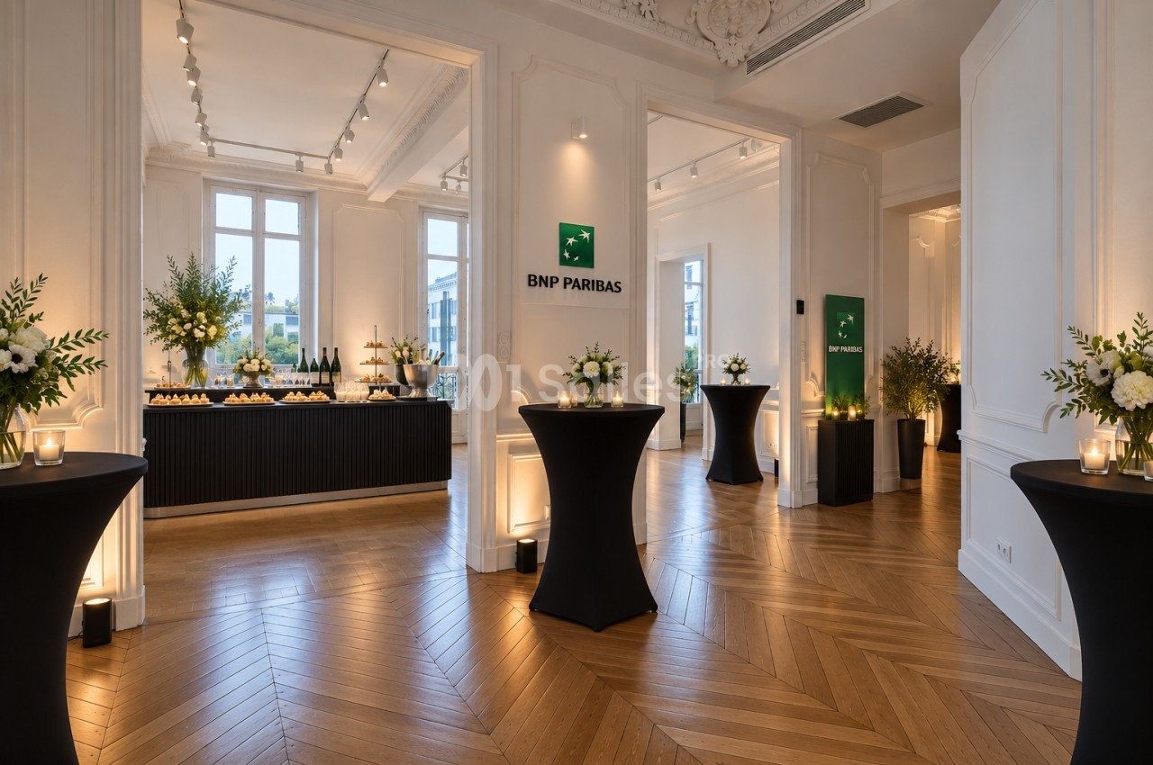 Salle de réception aux murs blancs moulurés et parquet en point de Hongrie, avec des tables hautes habillées de noir, des bouquets blancs et un buffet de boissons et de pièces salées. Let me check length: roughly 170 characters, too long. Need ≤125