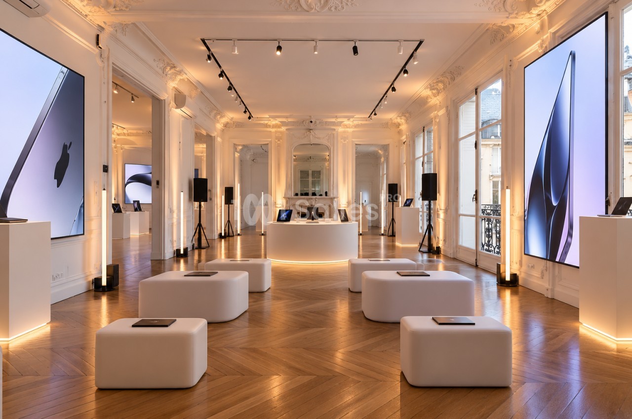 Grande salle au parquet en chevrons et moulures blanches, dotée de grands écrans lumineux sur les murs, de cubes blancs disposés au sol et d'un îlot central rond.