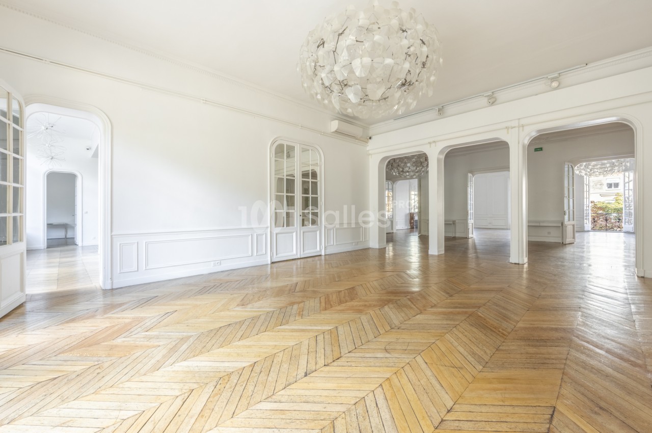 Grande pièce vide au parquet en chevrons, murs blancs à moulures, grand luminaire sphérique, enfilade de salles aux portes-fenêtres et arches.
