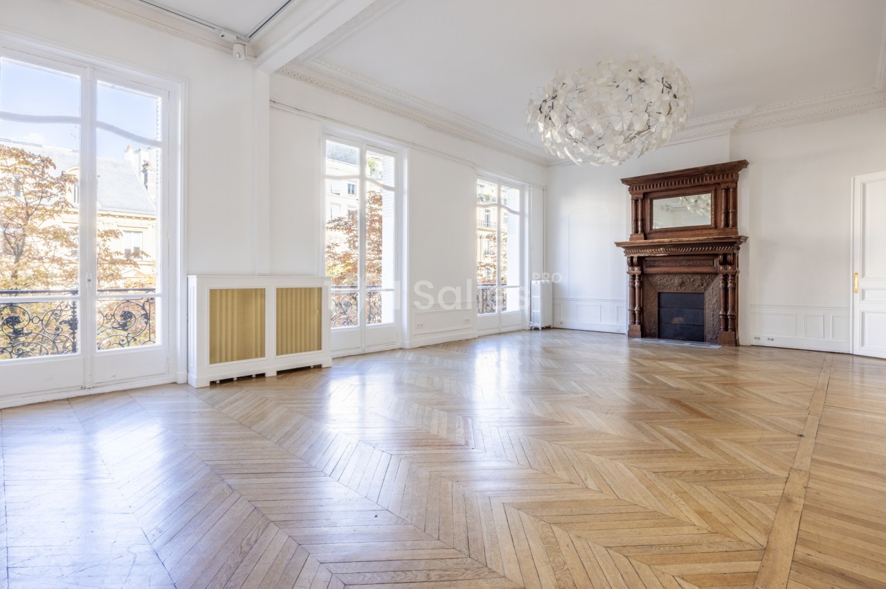Grande pièce vide au parquet en point de Hongrie, cheminée en bois sculpté ancienne, hautes fenêtres à balcons en fer forgé et lustre blanc.