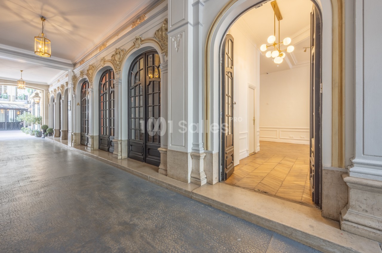 Galerie à arcades ornée de moulures dorées et de portes-fenêtres vitrées sombres, menant à une pièce claire au parquet en chevrons et à lustre à globes.