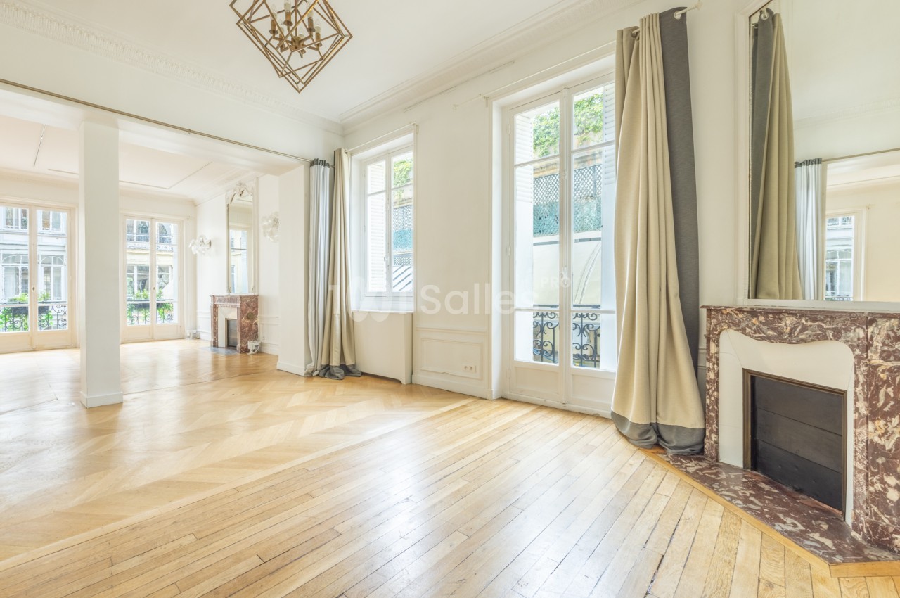 Grande pièce vide au parquet en chevrons clair, avec hautes portes-fenêtres, cheminées en marbre et rideaux beige, sans mobilier ni tables dressées.