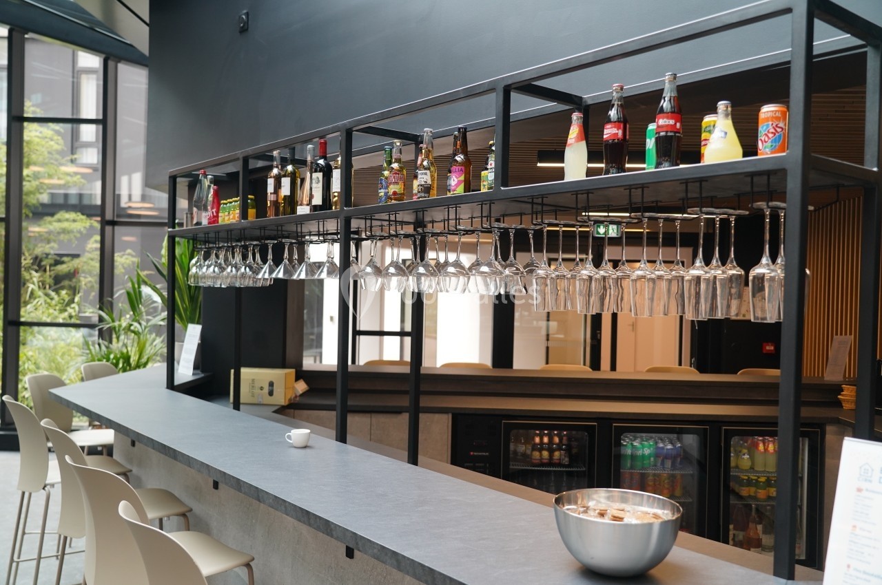 Bar moderne avec comptoir en pierre, verres suspendus, bouteilles alignées et vue sur un espace lumineux en arrière-plan.