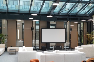 Salle lumineuse avec canapés, fauteuils, table basse, étagère de livres et écran mural, sur un sol en moquette grise.