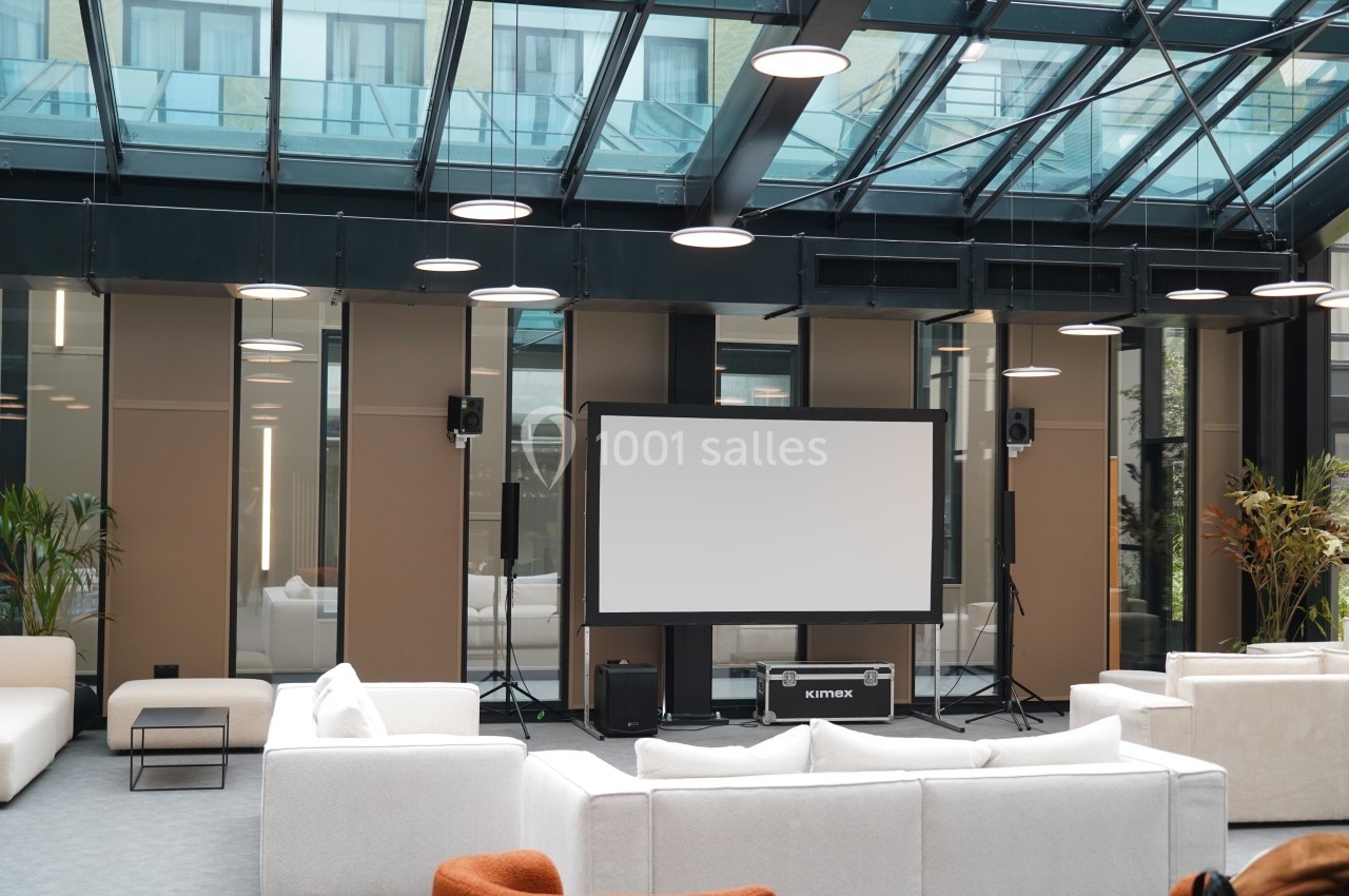 Salle moderne avec toit en verre, équipée d'un écran de projection, de canapés blancs et de plantes décoratives.