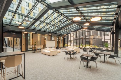 Salle lumineuse avec canapés, fauteuils, table basse, étagère de livres et écran mural, sur un sol en moquette grise.