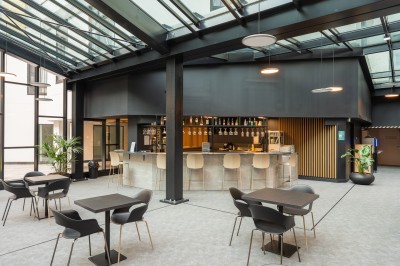 Salle lumineuse avec canapés, fauteuils, table basse, étagère de livres et écran mural, sur un sol en moquette grise.
