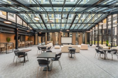 Salle lumineuse avec canapés, fauteuils, table basse, étagère de livres et écran mural, sur un sol en moquette grise.