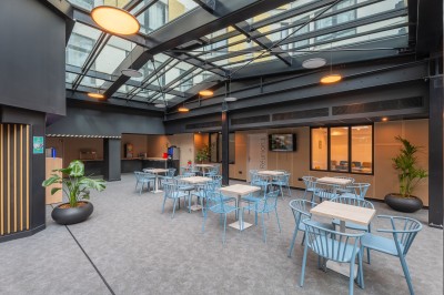 Salle lumineuse avec canapés, fauteuils, table basse, étagère de livres et écran mural, sur un sol en moquette grise.