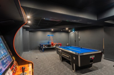 Salle lumineuse avec canapés, fauteuils, table basse, étagère de livres et écran mural, sur un sol en moquette grise.